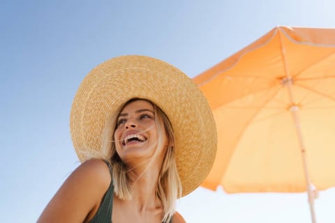 Woman wearing a straw hat smiling outdoors under an orange beach umbrella against a blue sky