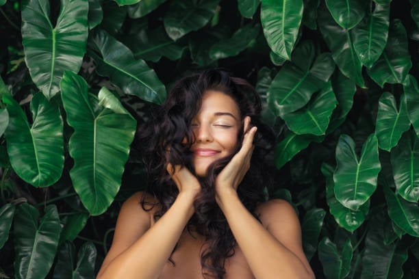 Woman with long dark wavy hair posing with hands to face against a backdrop of green tropical leaves