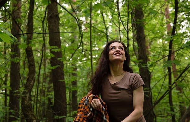 Woman with long dark hair wearing brown shirt gazing upward in dense green forest surrounded by tall trees
