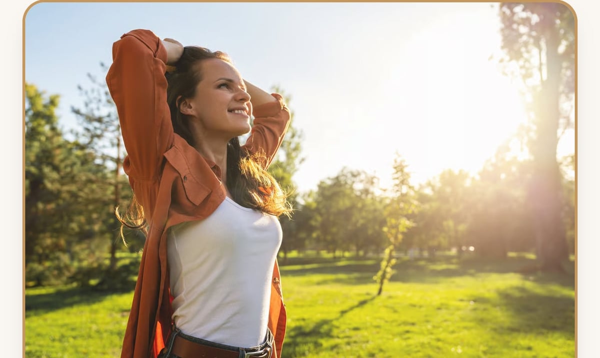 Woman smiling in sunny park wearing red jacket over white shirt, with lush green grass and trees in background