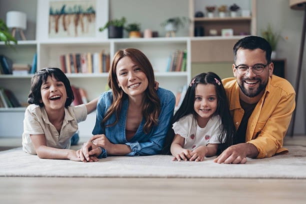Happy family of four smiling together at home, leaning on a table with bookshelves in the background