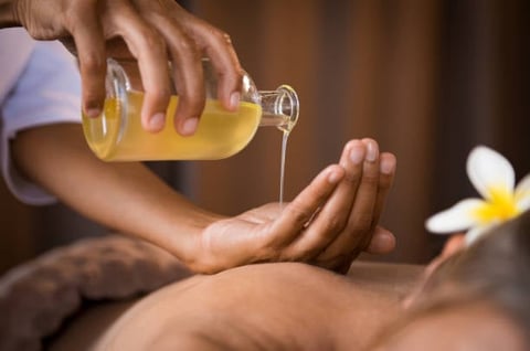 Therapist pouring golden massage oil onto clients back during spa treatment with white flower nearby