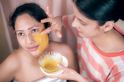Two women applying a golden face mask, with one holding a bowl of the treatment product during a skincare routine